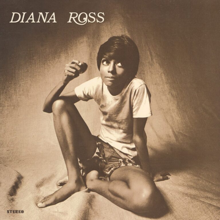 Diana Ross sitzt in lässiger Pose auf einem Stoffuntergrund und hält einen Apfel in der Hand.