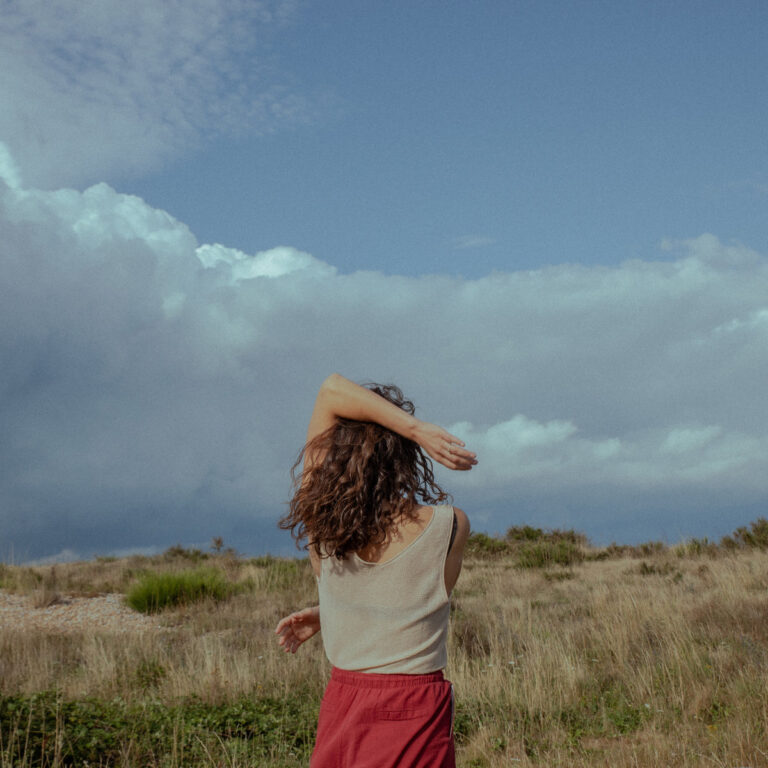 Eine Frau mit lockigem Haar, rotem Rock und beigem Top steht mit dem Rücken zur Kamera auf einer weiten Wiese unter dramatischem Himmel – Cover von I Still Want To Share.