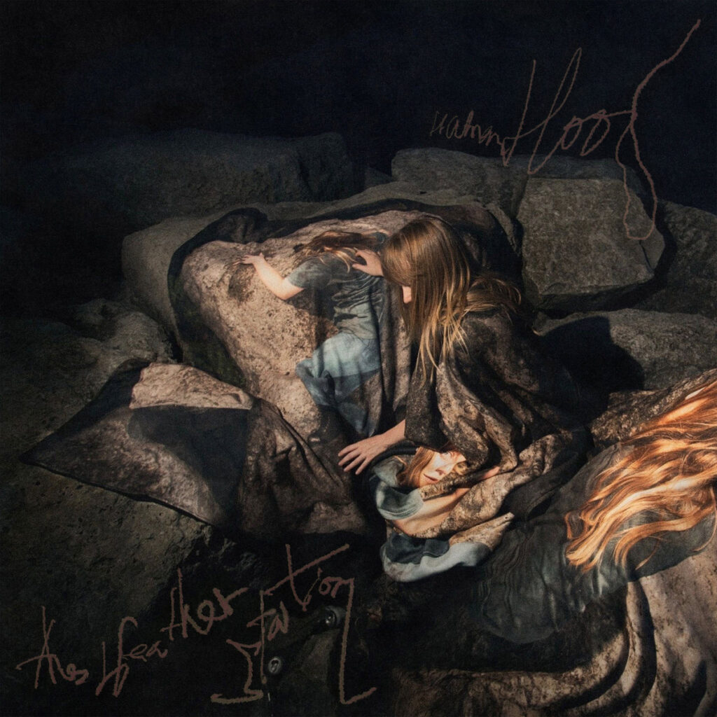Eine Frau liegt inmitten von Felsen, eingehüllt in dunkle Stoffe mit fotorealistischen Körperabbildungen – das Cover zu „Humanhood“ wirkt surreal, verletzlich und symbolträchtig.
