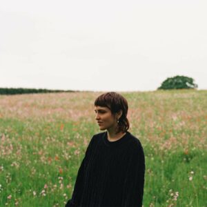Das Cover zeigt Dana Gavanski in einem dunklen Strickpullover auf einer weiten Blumenwiese unter grauem Himmel – ein ruhiges, nachdenkliches Porträt zwischen Natur, Melancholie und innerer Reise.