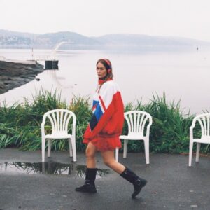 Anna of the North geht in rotem Kleid und schwarzer Jacke an weißen Plastikstühlen vorbei, im Hintergrund ein nebliger Fjord – ein ruhiges, nostalgisches Coverbild voller norwegischer Atmosphäre.