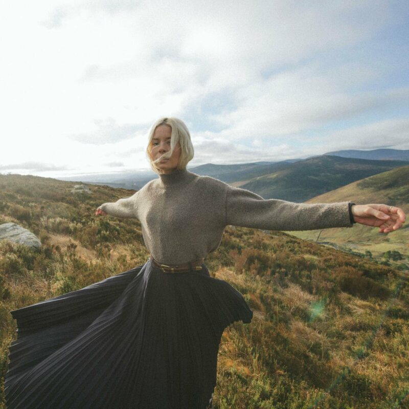 Nina Nesbitt steht mit ausgebreiteten Armen und wehender Kleidung inmitten schottischer Hügel – ein atmosphärisches Naturbild, das Freiheit und Verbundenheit zur Heimat ausstrahlt.
