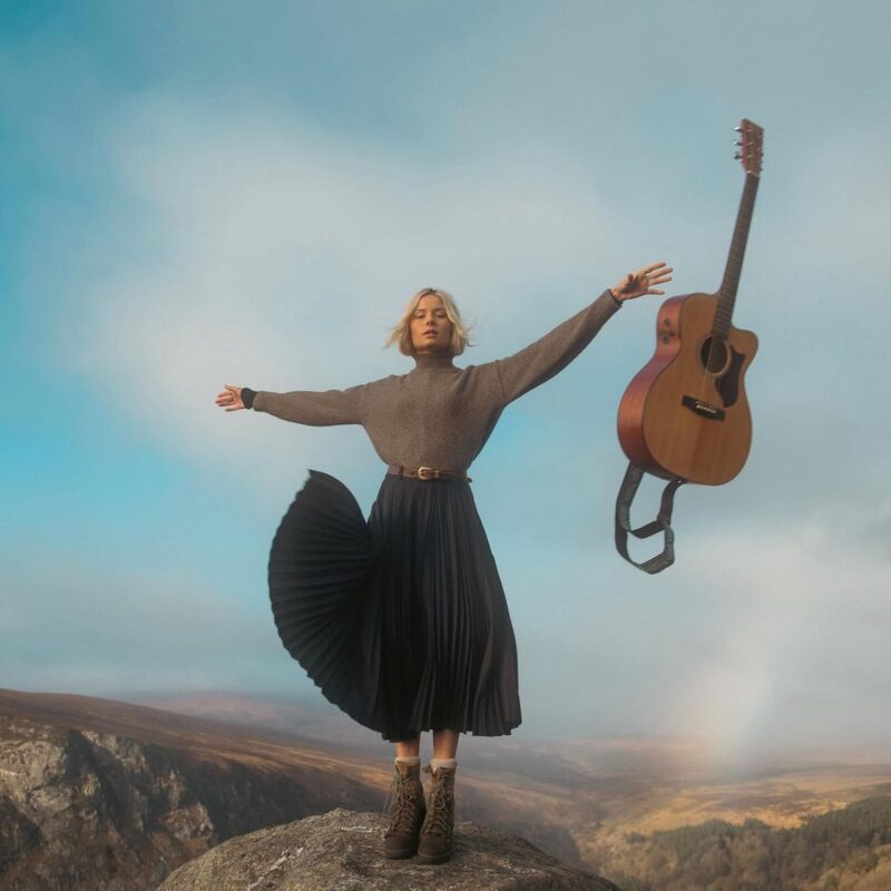 Cover von Mountain Music: Nina Nesbitt steht mit ausgestreckten Armen auf einem Felsen in wilder Landschaft, während eine Akustikgitarre in der Luft schwebt – frei, ruhig, symbolisch.