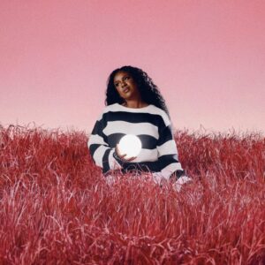 Nao sitzt in rot gefärbtem Gras unter rosa Himmel, trägt einen gestreiften Pullover und hält eine leuchtende Kugel – Cover zur Single „Light Years“.