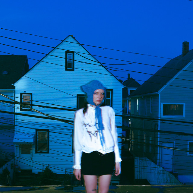 Ally Evenson steht vor blauen Häusern in der Dämmerung, umgeben von Stromleitungen – symbolträchtiges Artwork zur Dualität in ihrer Single „Scary/Calm“.