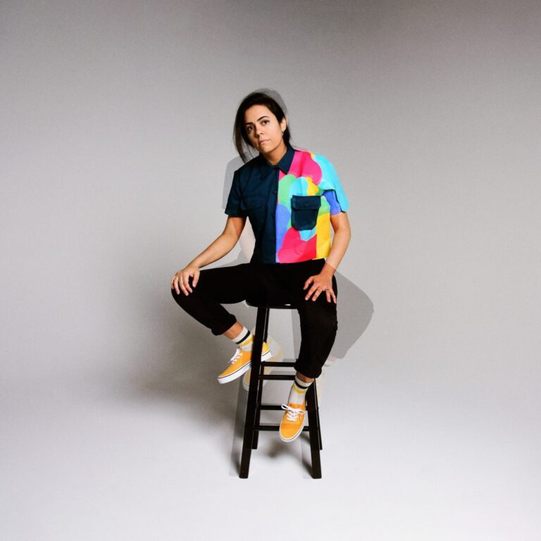 Sarah Tudzin alias Illuminati Hotties sitzt auf einem Hocker in schlichtem Studio-Setting. Ihr Hemd kombiniert dunkles Blau mit einer bunten, abstrakten Farbfläche – ein visuelles Statement für persönliche Dualität und kreative Freiheit.