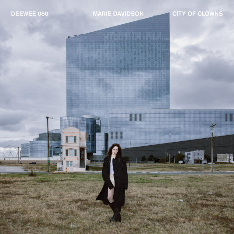 Das Cover von City of Clowns zeigt Marie Davidson in dunklem Mantel vor einer kalten, futuristisch wirkenden Glasarchitektur – ein Kontrast aus menschlicher Präsenz und urbaner Anonymität, der perfekt zur kritischen Klangästhetik des Albums passt.