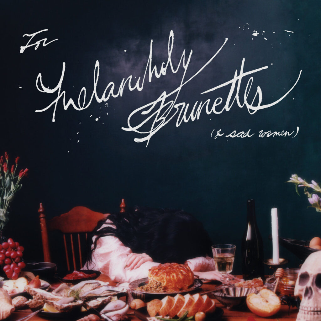 Albumcover von "For Melancholy Brunettes (& sad women)" von Japanese Breakfast: Michelle Zauner liegt mit dem Kopf auf einem überladenen Tisch mit Speisen, Blumen und einem Totenschädel – ein visuelles Stillleben der Melancholie.​