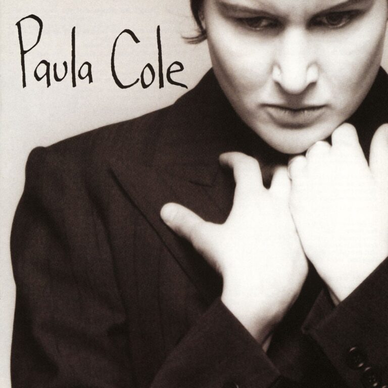 Paula Cole in einem dunklen Anzug mit verschränkten Armen vor neutralem Hintergrund.