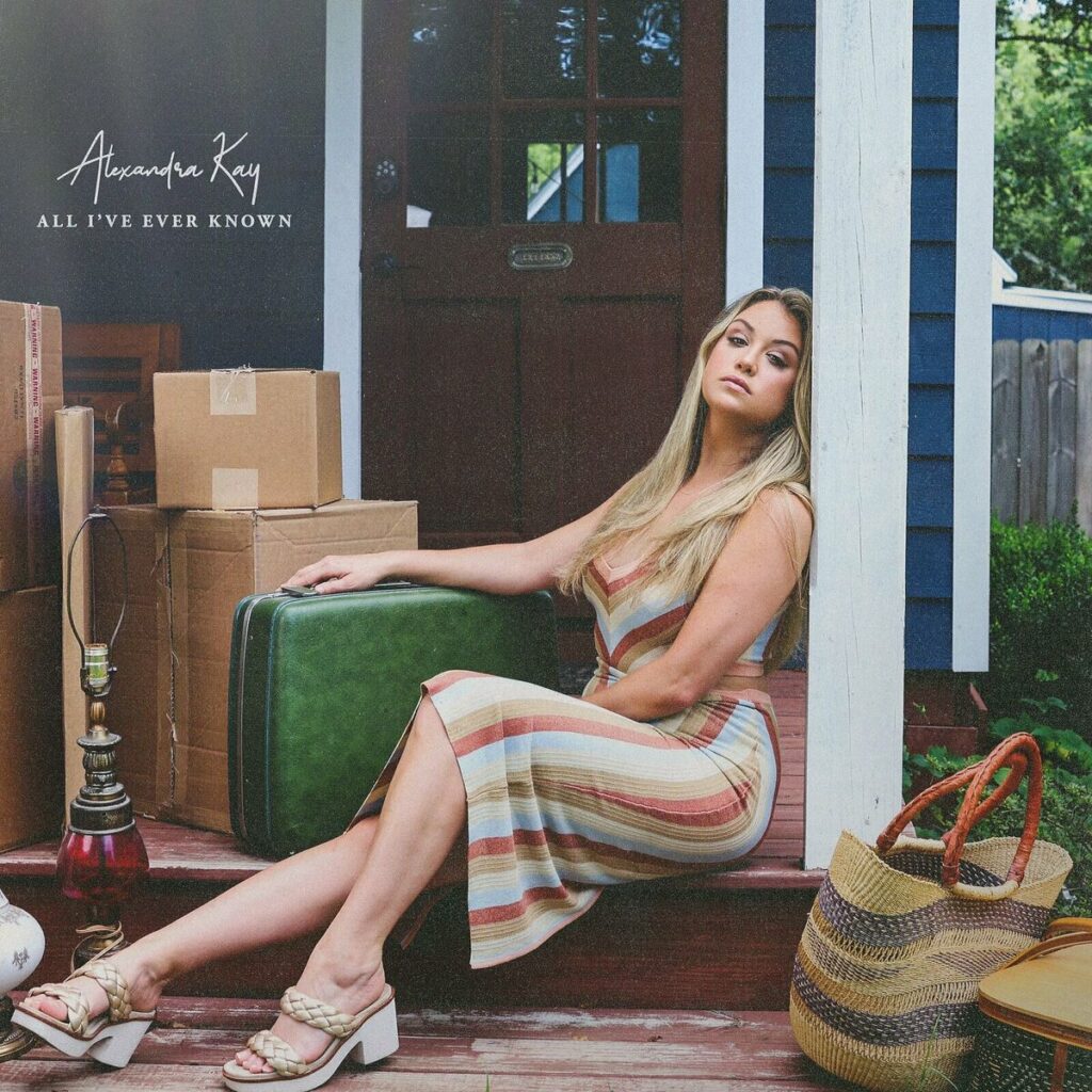 Albumcover von All I’ve Ever Known: Alexandra Kay sitzt in einem gestreiften Kleid mit Koffern und Umzugskartons auf einer Veranda, nachdenklich, aber entschlossen.