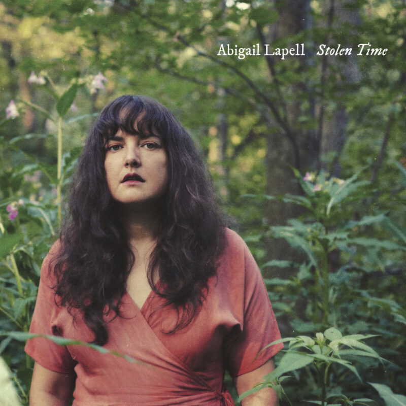 Abigail Lapell steht in dichtem Grün eines Waldes, trägt ein rosafarbenes Wickelkleid und blickt ernst ins Bild – Albumcover von „Stolen Time“.