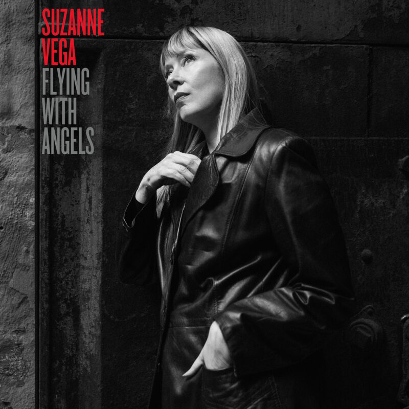 Schwarz-weiß-Fotografie von Suzanne Vega in Ledermantel, an eine dunkle Wand gelehnt, mit nachdenklichem Blick nach oben – das Cover zu „Flying With Angels“ strahlt Reife, Ernsthaftigkeit und zeitlose Eleganz aus.