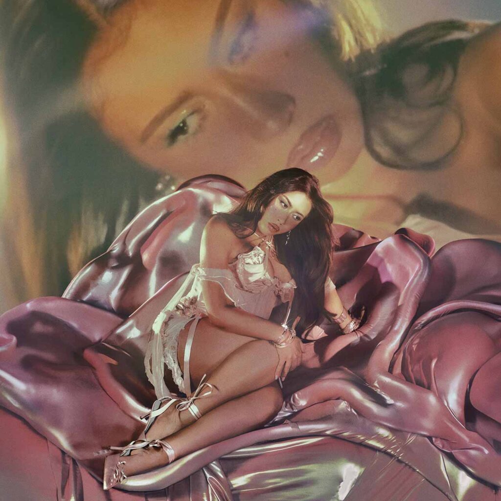 Kali Uchis sitzt auf einer glänzenden, rosafarbenen Rosenstruktur, gekleidet in ein transparentes, blütenartiges Kleid. Im Hintergrund erscheint ihr eigenes Gesicht übergroß und weichgezeichnet – ein ätherisches Porträt zwischen Sinnlichkeit, Verletzlichkeit und ikonischer Stärke. Die Szene erinnert an barocke Heiligenbilder und moderne Pop-Ästhetik zugleich.