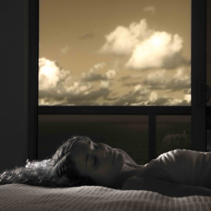 Frau liegt verträumt auf dem Bett, während draußen ein dramatischer Himmel im Fenster erscheint – Stimmung zwischen Intimität und Melancholie.