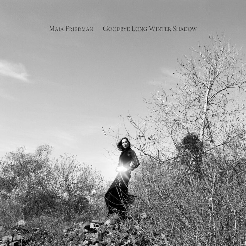 Maia Friedman steht in winterlicher Landschaft, in Schwarzweiß, von Sonnenlicht durchbrochen – Albumcover Goodbye Long Winter Shadow
