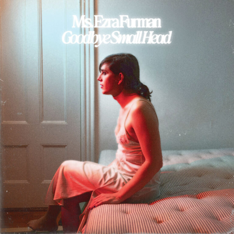 Ezra Furman sitzt im weißen Nachthemd auf einer Matratze, umgeben von kühlem und warmem Licht – ein Moment zwischen Rückzug und Aufbruch.