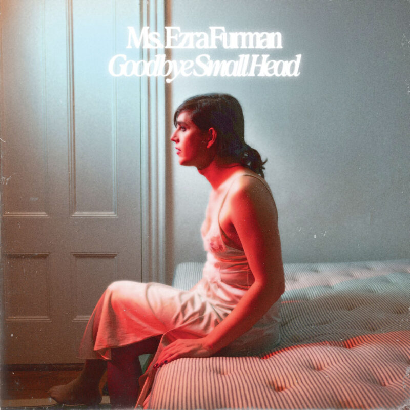 Ezra Furman sitzt im weißen Nachthemd auf einer Matratze, umgeben von kühlem und warmem Licht – ein Moment zwischen Rückzug und Aufbruch.
