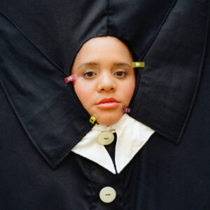 Lido Pimienta mit eindringlichem Blick in avantgardistischer schwarzer Stoffskulptur, eingefasst von bunten Klammern