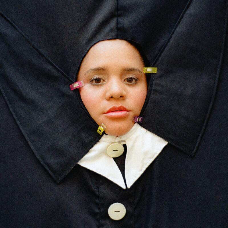 Lido Pimienta mit eindringlichem Blick in avantgardistischer schwarzer Stoffskulptur, eingefasst von bunten Klammern