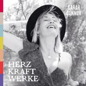 Albumcover von Herz Kraft Werke mit Sarah Connor lachend im Sonnenlicht mit Hut und Tattoos, links farbige Balken.