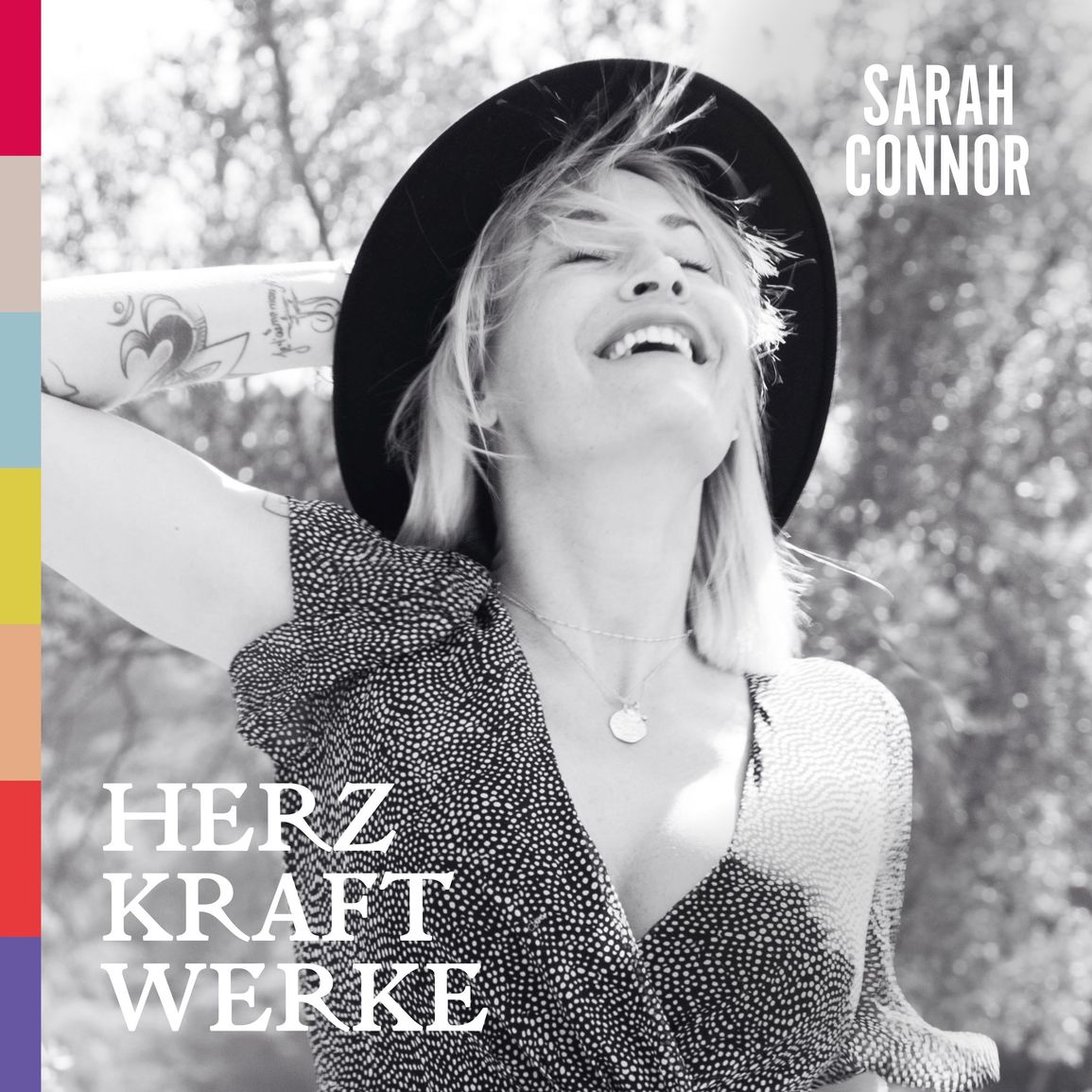Albumcover von Herz Kraft Werke mit Sarah Connor lachend im Sonnenlicht mit Hut und Tattoos, links farbige Balken.