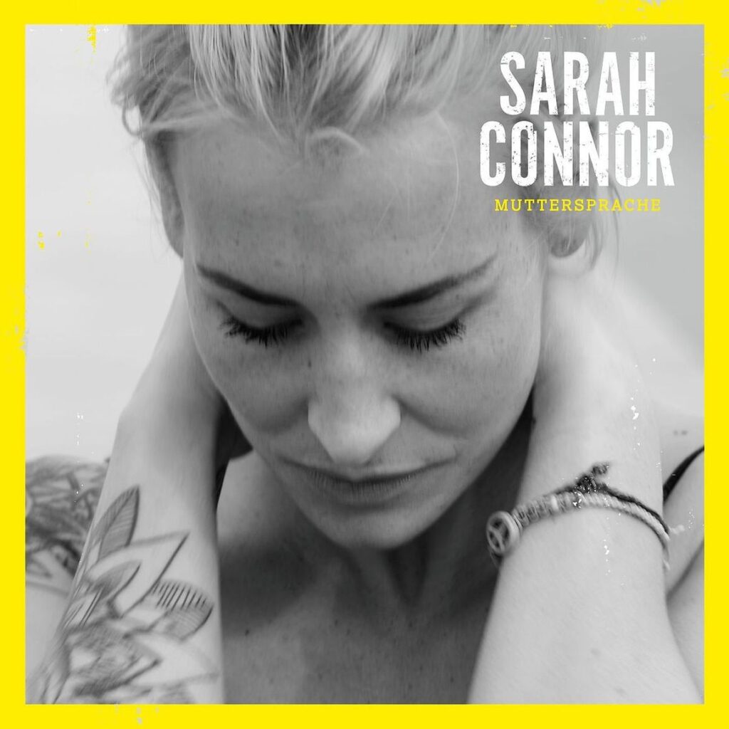 Schwarzweißporträt von Sarah Connor mit gesenktem Blick und Tattoo, eingerahmt von einem leuchtenden Gelb