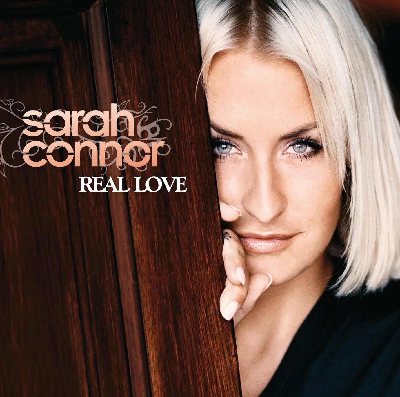Sarah Connor lehnt an hölzernem Türrahmen und blickt direkt in die Kamera – Cover ihres Albums „Real Love“