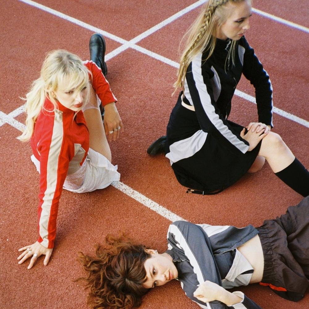 Die drei Mitglieder der Band Blusher posieren in sportlichen Outfits auf einer roten Laufbahn – ein stilisiertes Pressefoto zur Single „WHATEVERWHATEVER“, das Energie, Retro-Vibes und emotionale Unabhängigkeit visuell zusammenbringt.