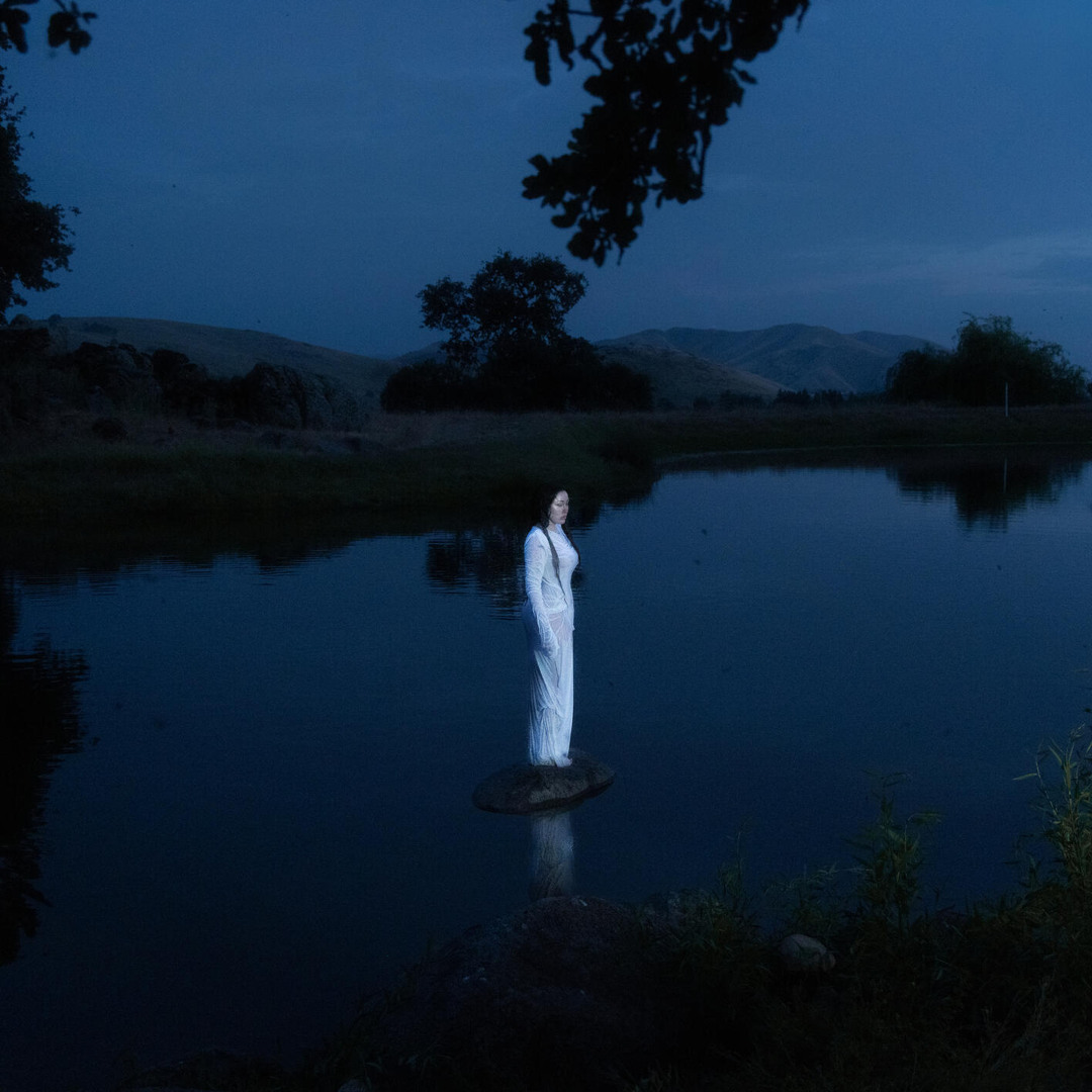 Noah Cyrus in weißem Kleid bei Dämmerung auf einem Stein im Wasser stehend, umgeben von dunkler Natur und Spiegelungen – Cover von „I Saw the Mountains“.