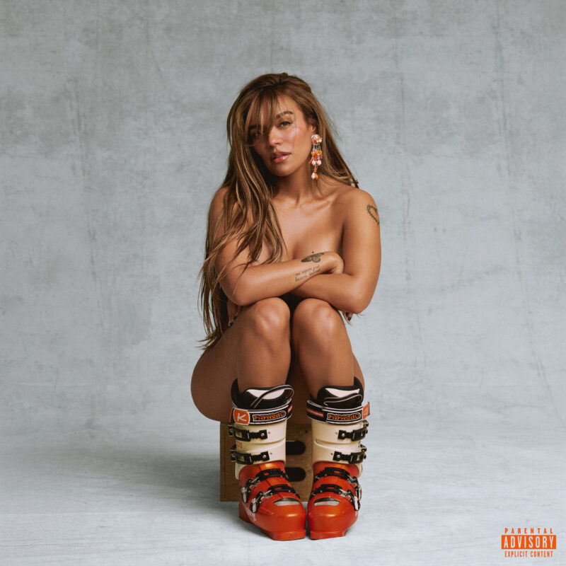 Karol G sitzt nackt in Skischuhen auf einem Holzhocker vor grauem Hintergrund – Cover zur Single LATINA FOREVA.