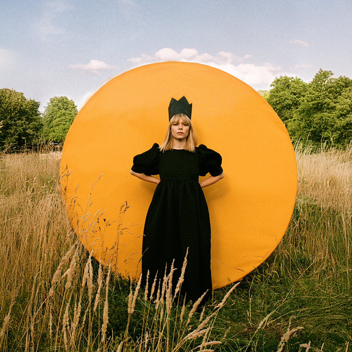 Chloe Foy steht mit Papierkrone vor einer gelben Kreisscheibe in einem Feld – Albumcover Complete Fool