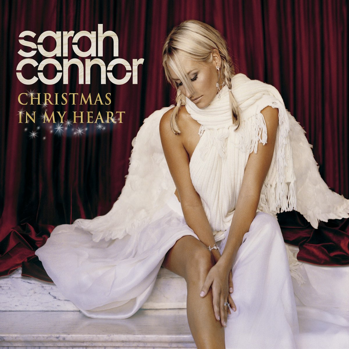 Sarah Connor sitzt als geflügelter Engel in Weiß vor rotem Samtvorhang – Albumcover „Christmas In My Heart“.