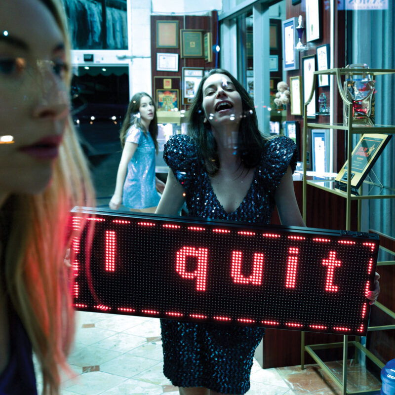 Frau in glitzerndem Kleid hält Leuchtschild mit „I Quit“ in einem retro Dekor-Shop mit Auszeichnungen