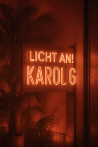 Ein leuchtendes Neon-Schild mit der Aufschrift „LICHT AN! KAROL G“ in warmem Kupfer-Orange hängt vor einer Wand mit tropischen Palmen im Hintergrund. Das Licht wirft weiche Reflexe auf die Umgebung und erzeugt eine stimmungsvolle, glamourös-lateinamerikanisch inspirierte Atmosphäre.