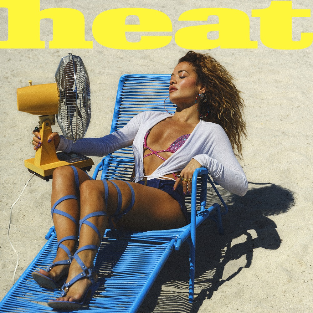 Rita Ora liegt auf einer blauen Liege mit gelbem Ventilator am Strand – Retro-Look, Pop-Attitüde und Hitzegefühl pur für ihre Single „Heat“