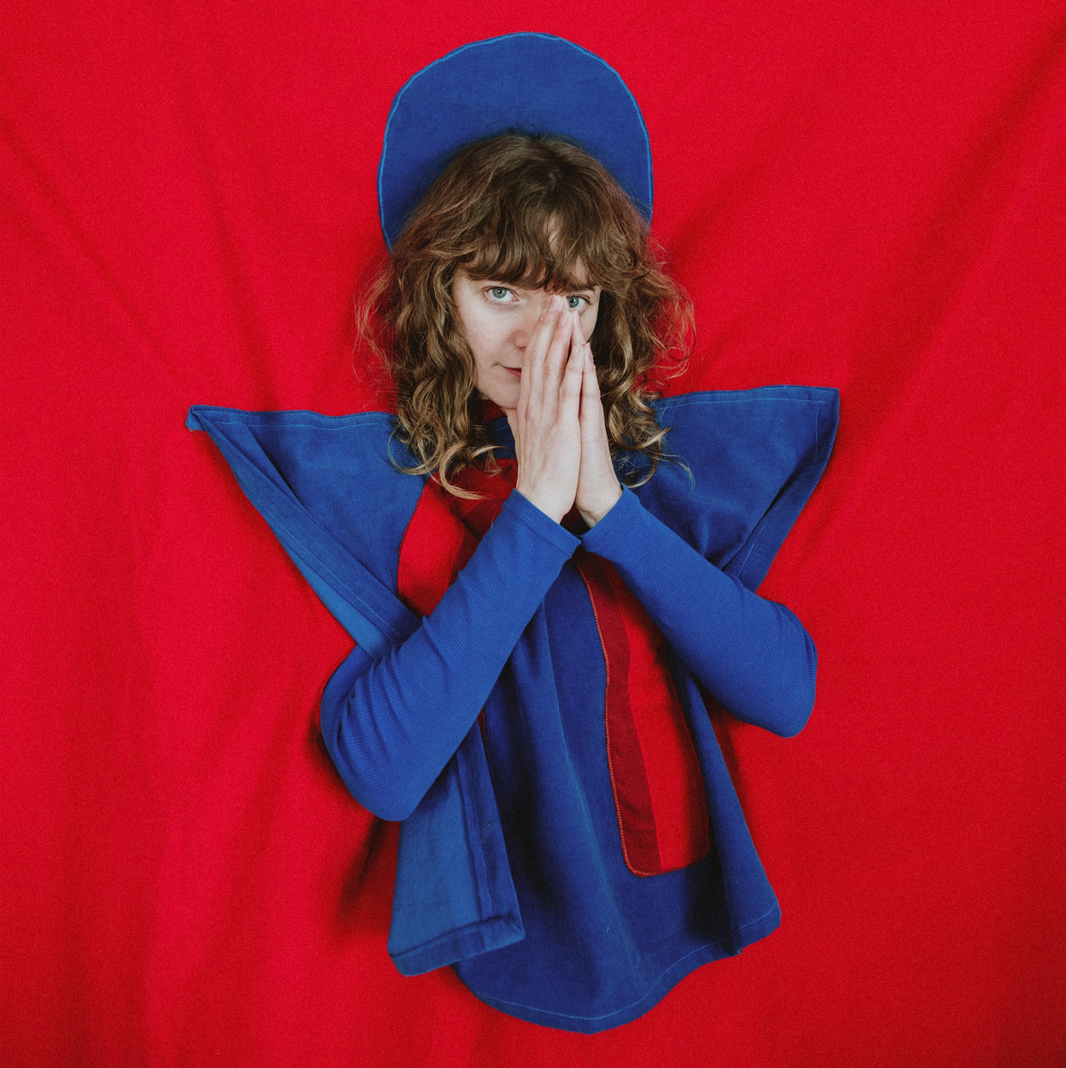 Künstlerin Me Lost Me vor rotem Stoff, in futuristisch-blauem Gewand – poetisches Albumcover zu „This Material Moment“.