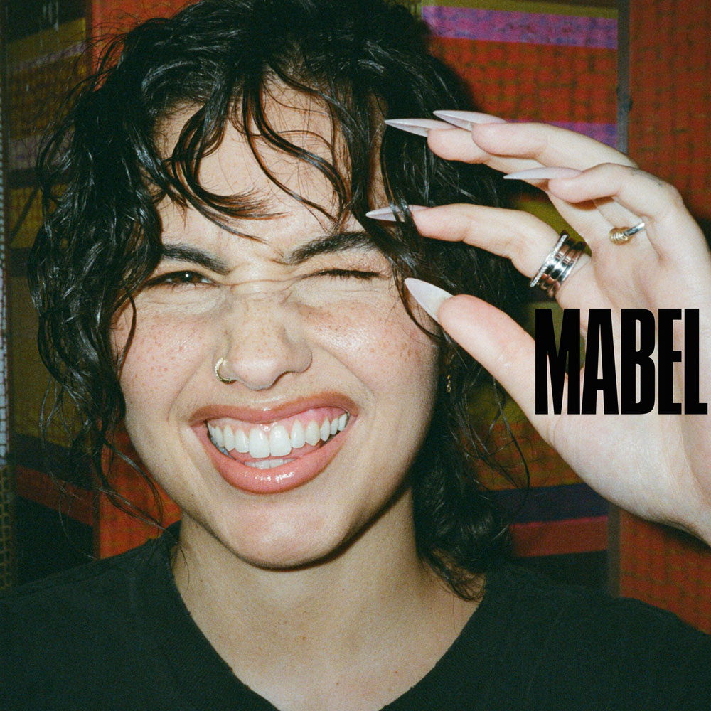 Nahaufnahme von Mabel mit breitem Lächeln und hochgehaltener Hand, verspielter DIY-Vibe. Hover-Zeile: Mabel grinst in die Linse wie ins Chaos – und nimmt uns mit auf ihr wildestes Mixtape bisher.