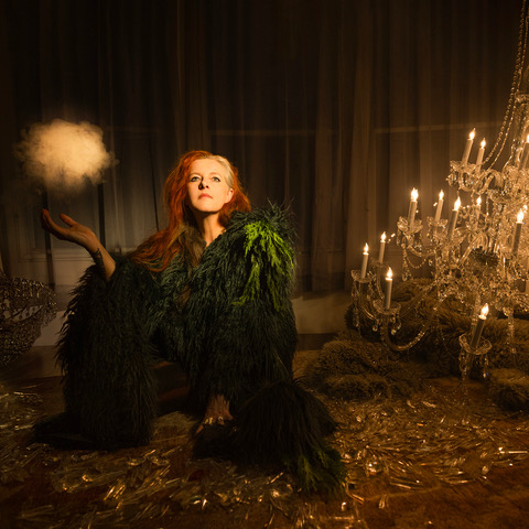 Neko Case in grünem Federkleid vor Kronleuchtern, mystisch inszeniert für das Album „Neon Grey Midnight Green“