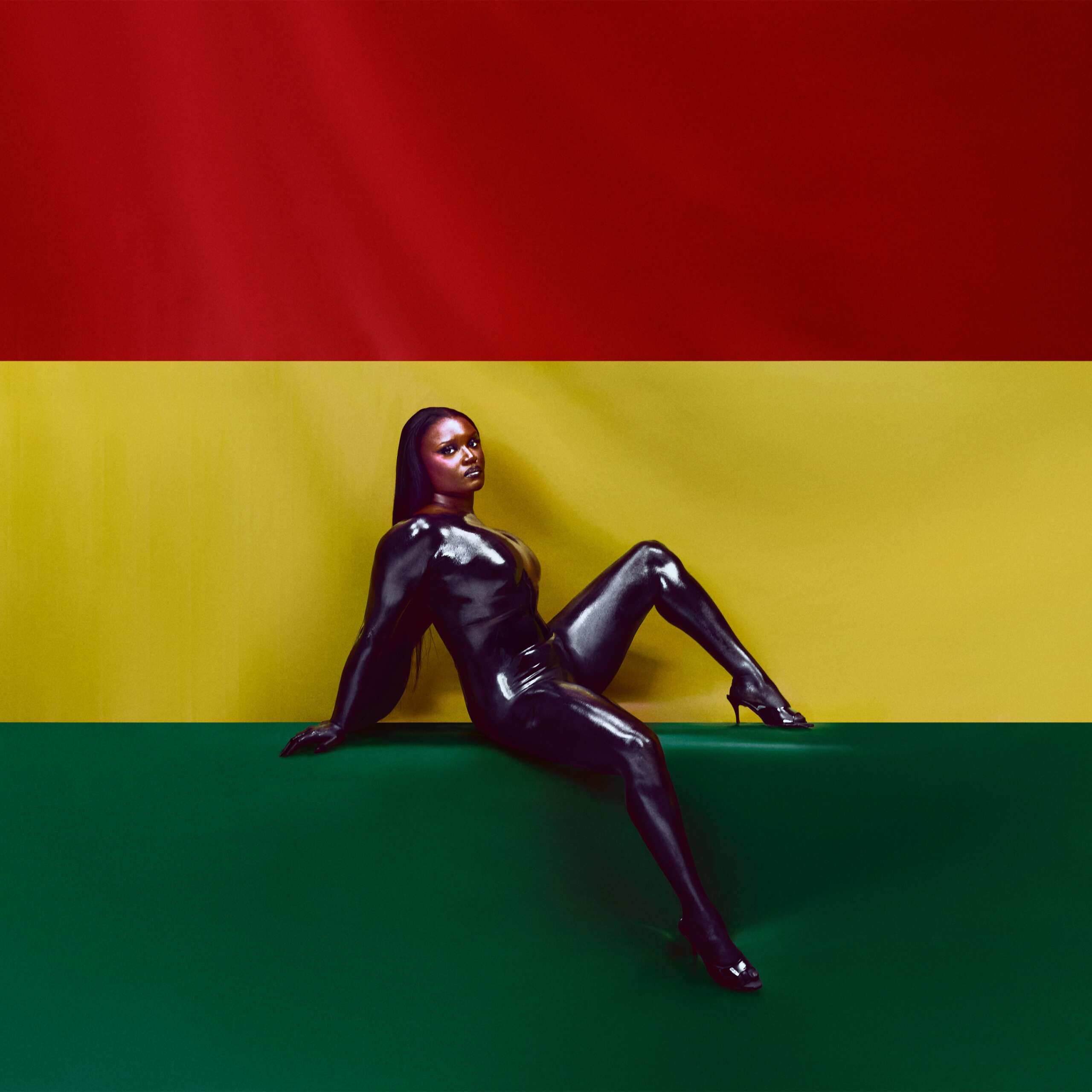 Amaarae lehnt in schwarzem Latexanzug vor einem Hintergrund in den Farben Rot, Gelb und Grün – stilisierte Referenz an die ghanaische Flagge.