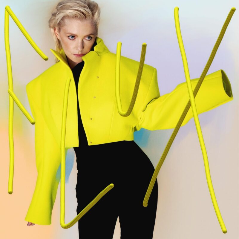 Albumcover von Alison Goldfrapps Flux mit der Sängerin in leuchtend gelber Jacke vor pastellfarbenem Hintergrund.