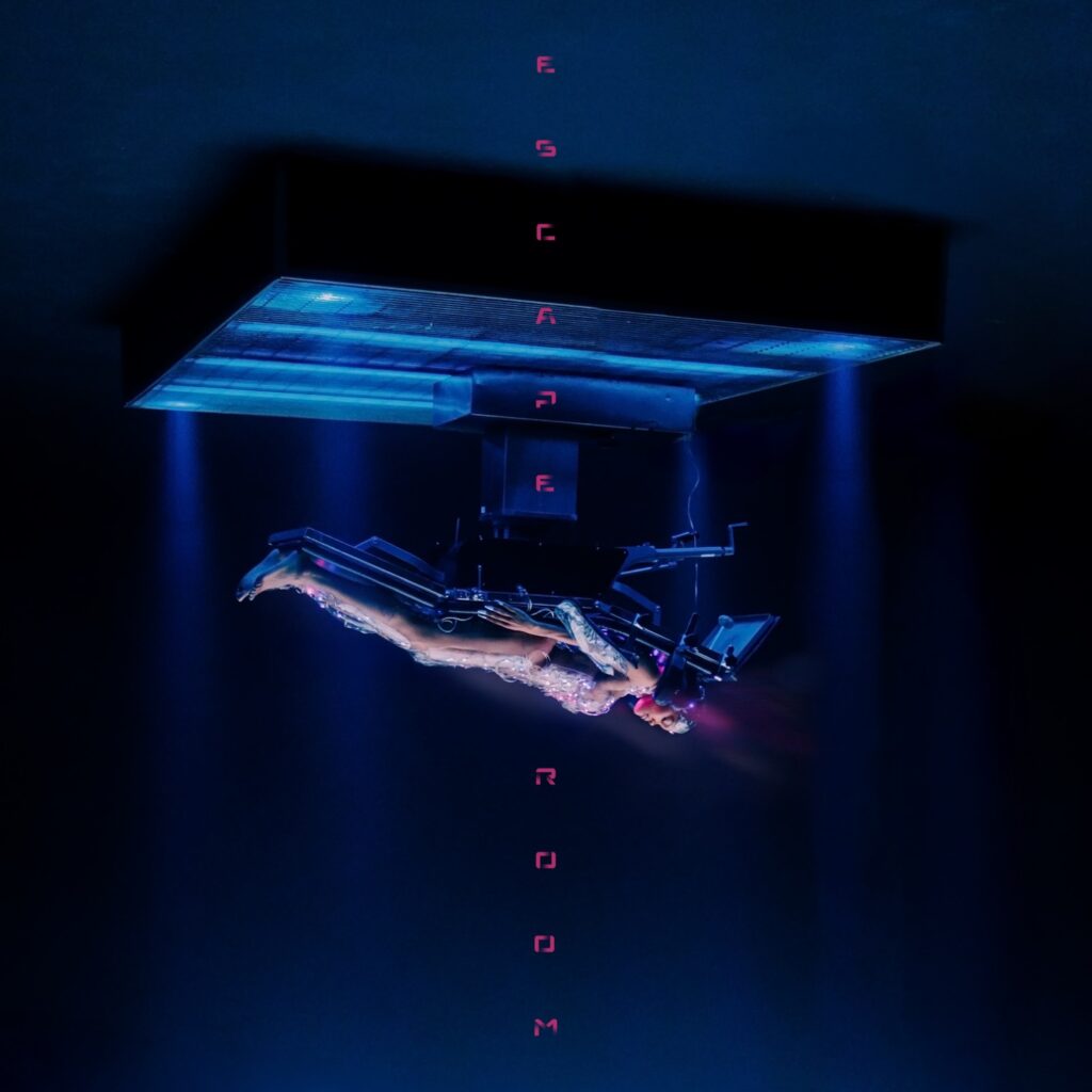 Albumcover mit futuristischer Szene: ein nackter Körper liegt auf einem umgedrehten Operationstisch, umgeben von kaltem blauen Licht und schwebender Typografie „ESCAPE ROOM“.