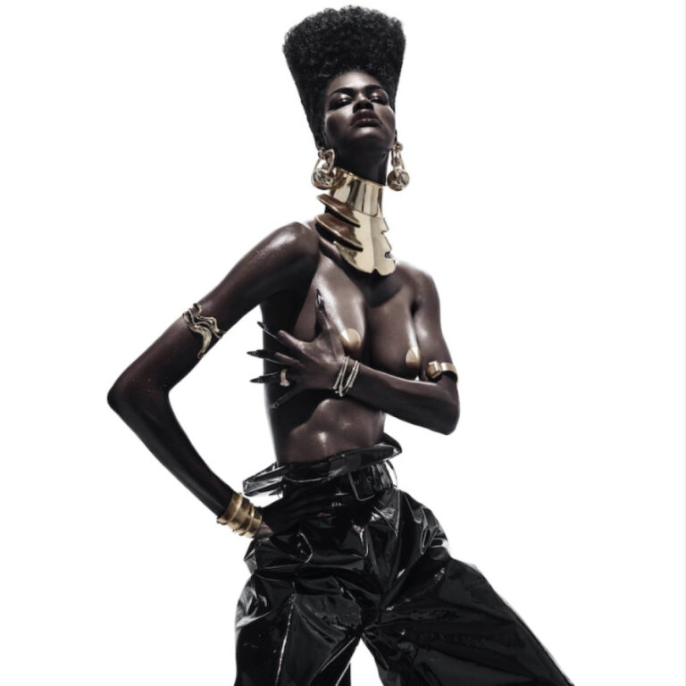 Hochglänzendes Studio-Porträt: Teyana Taylor topless mit goldener Statement-Halskrause und Schmuck, hochgestecktem Afro, schwarzen Vinyl-Hosen, weißer Hintergrund, kraftvolle Pose.