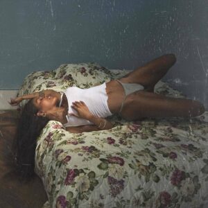 Quadratisches Albumcover: Teyana Taylor in weißem Tanktop auf einem geblümten Bett, halb liegend in einem düsteren, blaugrünen Zimmer; körnige, intime Lichtstimmung.