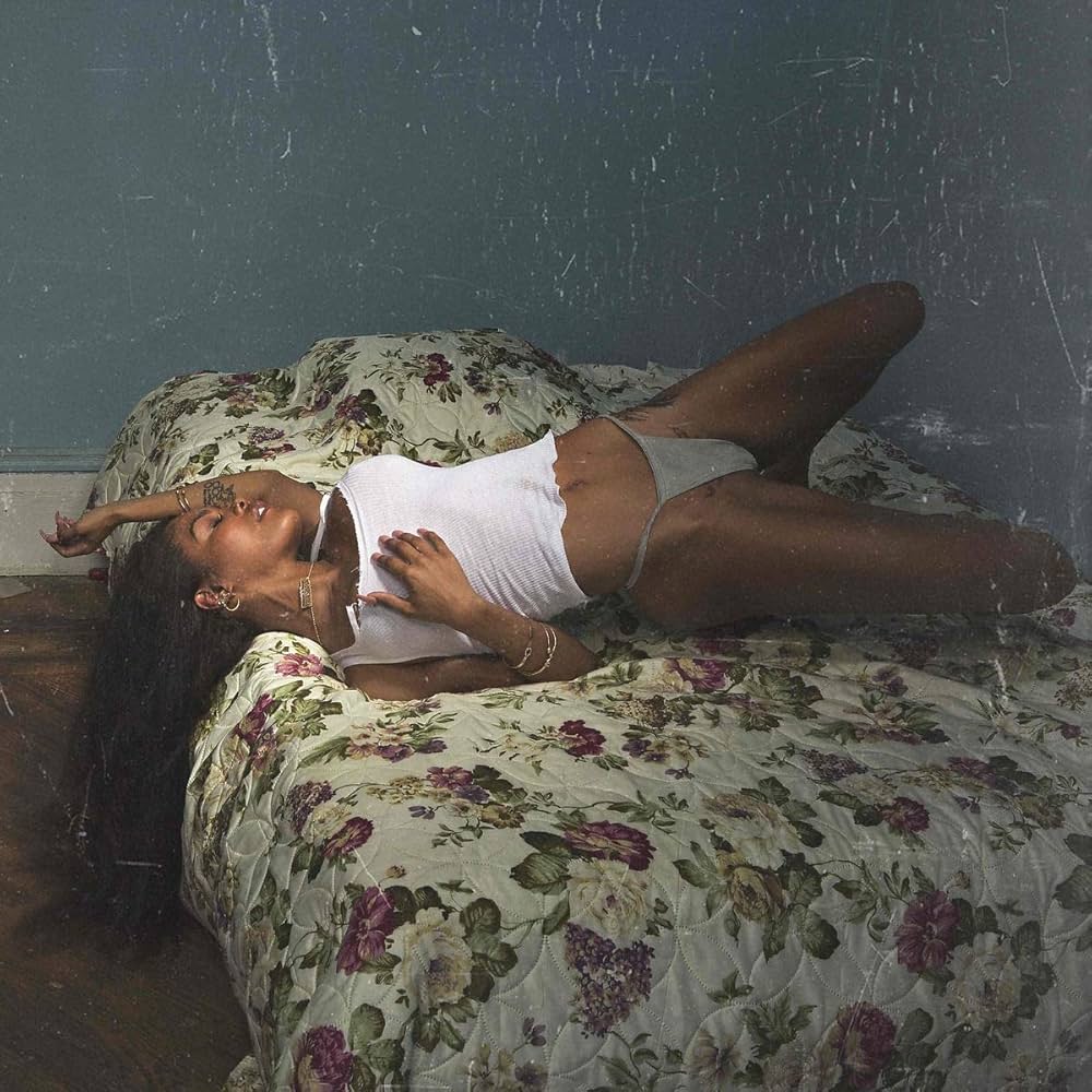 Quadratisches Albumcover: Teyana Taylor in weißem Tanktop auf einem geblümten Bett, halb liegend in einem düsteren, blaugrünen Zimmer; körnige, intime Lichtstimmung.