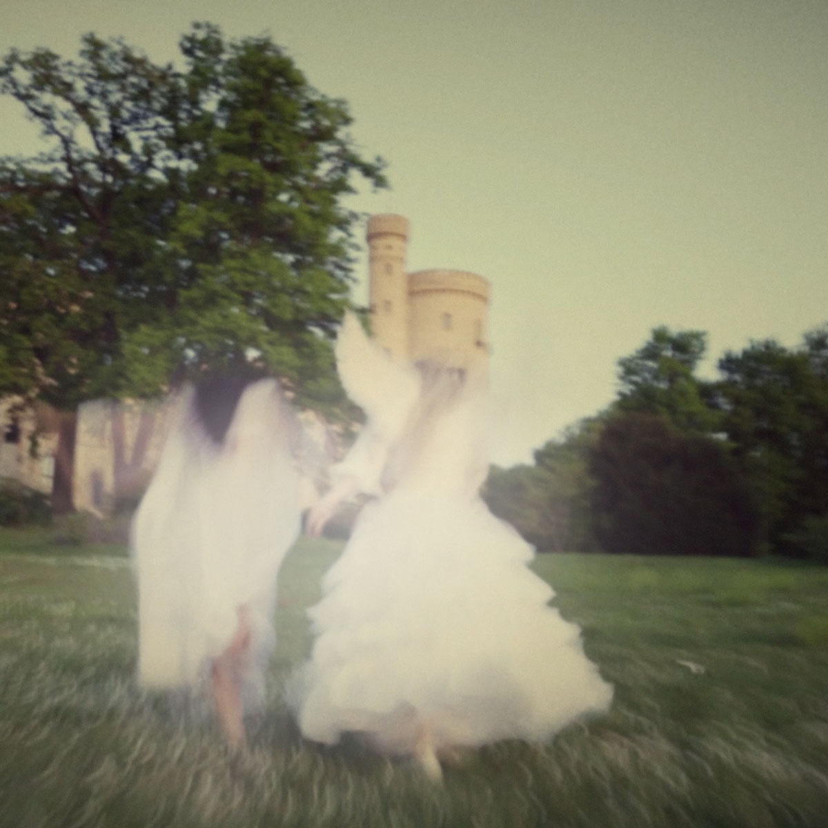 Verschwommenes Foto von zwei Frauen in weißen Kleidern, die Hand in Hand über eine Wiese auf ein Schloss zulaufen, im Hintergrund grüne Bäume.
