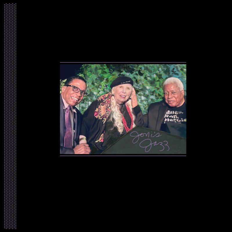 Fotografie mit Joni Mitchell in schwarzer Baskenmütze und besticktem Cape zwischen Herbie Hancock und Wayne Shorter, vor grünem Blätterhintergrund; unten handschriftlich „Joni’s Jazz“.
