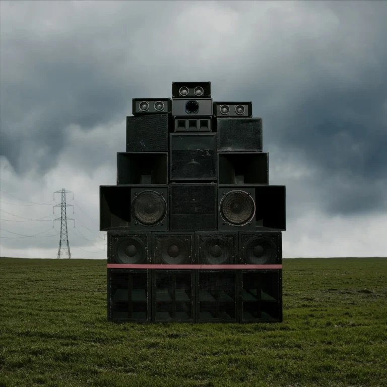 Ein turmartiges Stack aus schwarzen Lautsprechern steht auf einer grünen Wiese unter dunklem Wolkenhimmel; im Hintergrund ein Strommast, das Bild wirkt rau und monumental.