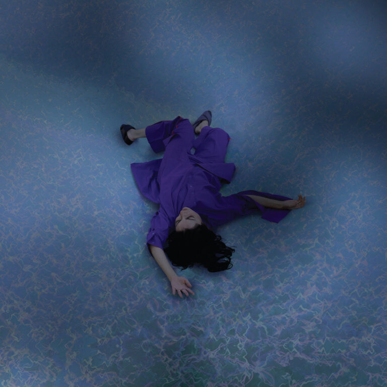 Aus der Vogelperspektive liegt eine Person in violettem Outfit auf einer blau schimmernden, wasserartig gemusterten Fläche, Arme und Beine locker ausgestreckt, weiches Licht, viel negative Fläche.