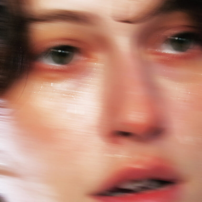 Unscharfes Nahporträt eines Gesichts mit grünlichen Augen und weichen Zügen, verschwommen in horizontalen Bewegungen, zwischen Nähe und Entrückung.