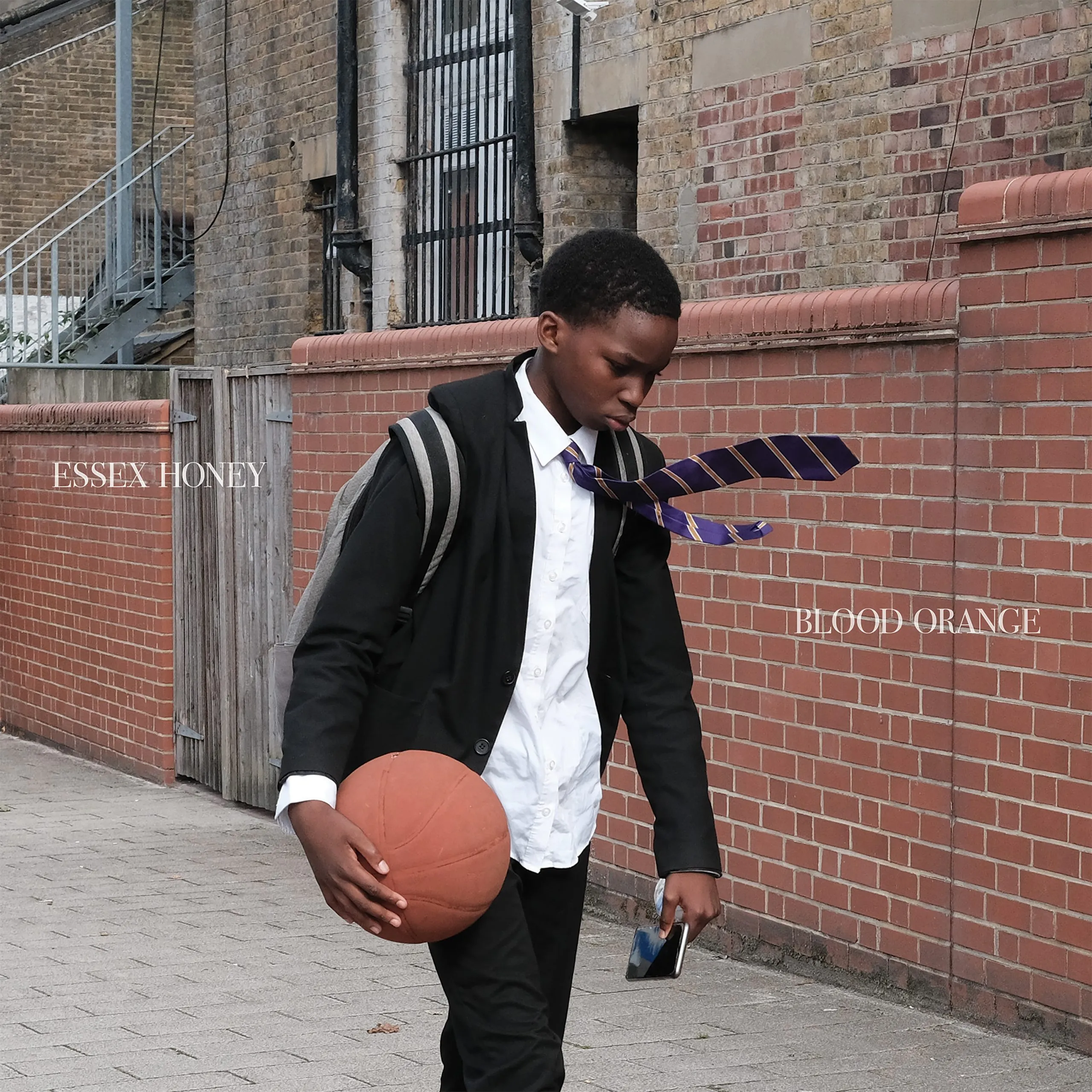 Ein Jugendlicher in Schuluniform geht mit gesenktem Blick an einer Backsteinmauer entlang, hält einen Basketball und ein Handy, die Krawatte weht im Wind.